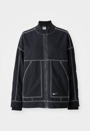 Veste noire avec un col montant, fermeture éclair, couture blanche contrastante et poches latérales. Dispose d'un petit logo Nike en bas à droite.