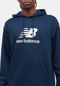 Osoba nosící tmavě modrou mikinu s bílým logem a nápisem New Balance na hrudi, čelem k nám s viditelnými rameny a krkem.