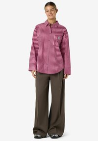 Red and white striped shirt with long sleeves, button front, and a chest pocket, paired with brown wide-leg trousers. Gray sneakers are visible.