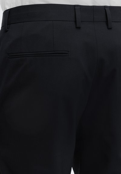 Gros plan de pantalon noir de costume vu de dos, montrant une poche passepoilée simple, des passants de ceinture et une partie de la ceinture d'une chemise blanche.