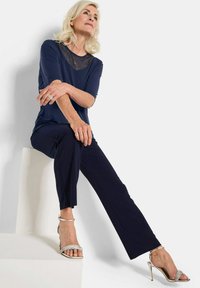 Navy-Oberteil mit einem durchsichtigen gepunkteten Ausschnitt, kombiniert mit navyfarbenen schmalen Hosen. Das Model trägt metallische Riemchen-Heels und sitzt auf einer weißen Plattform.