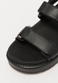Black leather sandal featuring dual adjustable straps with gold hardware, a textured footbed, and a sturdy, slightly raised sole.