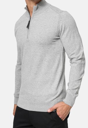 Mann trägt einen hellgrauen Pullover mit Viertel-Reißverschluss und langen Ärmeln sowie schwarze Hose, Hand in der Tasche, vor einfarbigem Hintergrund.