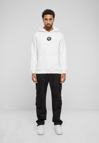 Felpa bianca con cappuccio e un logo circolare nero, abbinata a pantaloni cargo neri con numerose tasche. Indossata con sneaker bianche.