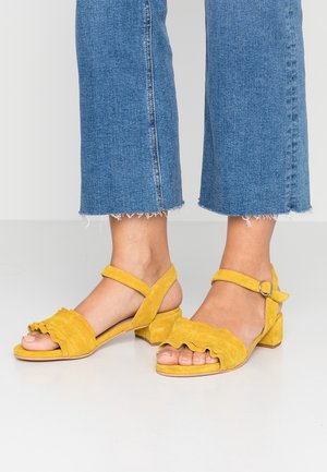 Pieds portant des sandales en daim jaune avec des brides froufroutantes et une boucle à la cheville, associées à un jean bleu court à ourlet effiloché.