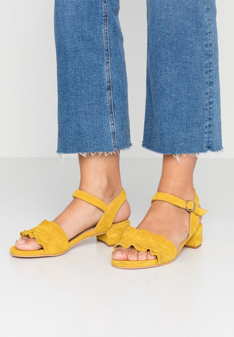Pieds portant des sandales en daim jaune avec des brides froufroutantes et une boucle à la cheville, associées à un jean bleu court à ourlet effiloché.