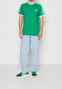 Grön bomullströja med vita accenter och Adidas-logotyp, matchad med ljusblå vida jeans och gröna mockaskor med vita ränder.