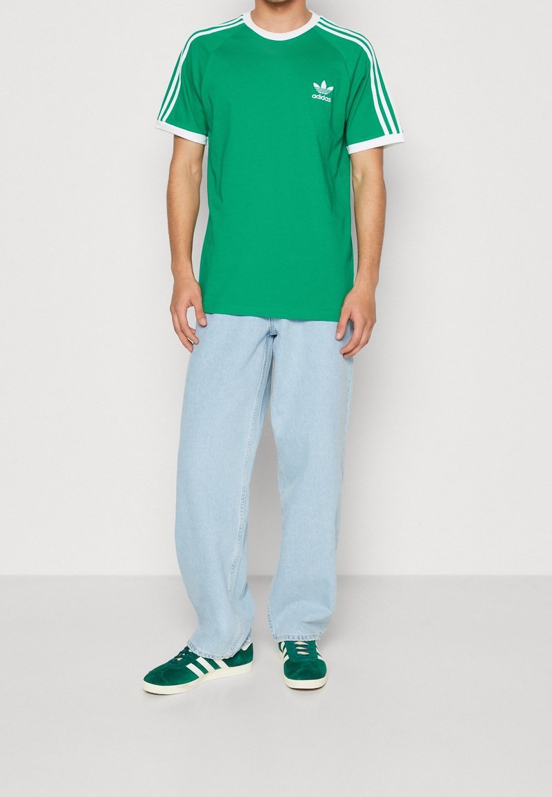 Grön bomullströja med vita accenter och Adidas-logotyp, matchad med ljusblå vida jeans och gröna mockaskor med vita ränder.