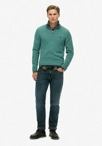 Sweter w kolorze morskim z dzianiny z zamkiem pod szyją, zestawiony z koszulą w kratę, niebieskimi dżinsami i czarnymi butami o fakturowanej powierzchni oraz podwiniętymi mankietami.