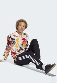 Camisa polo de manga longa com estampado gráfico colorido, colarinho branco, combinado com calças de treino pretas com riscas brancas nas laterais. Ténis pretos.
