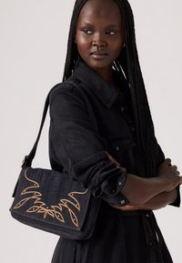 Sac à épaule en denim foncé avec broderie dorée dans un motif floral, comportant une fermeture à rabat et une bandoulière ajustable. Le sac a une finition texturée.