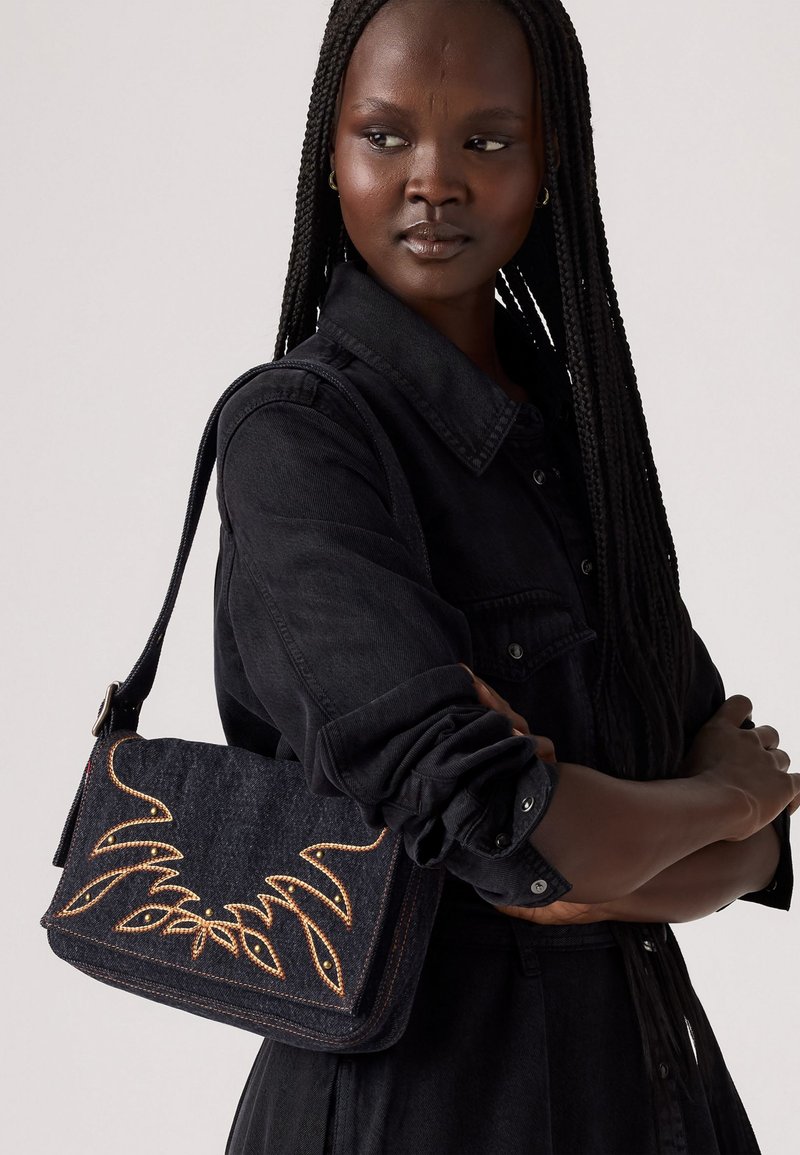 Sac à épaule en denim foncé avec broderie dorée dans un motif floral, comportant une fermeture à rabat et une bandoulière ajustable. Le sac a une finition texturée.