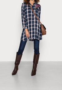 Mörkblå rutig skjorttunica med långa ärmar, kombinerad med mörka jeans och bruna texturerade knähöga stövlar. En brun axelväska bärs.