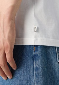 Hvid bomulds t-shirt med et side-mærke, parret med blå denim jeans. Hånden hviler på t-shirtens kant, som viser den bløde stoftekstur.
