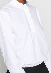 Vit bomullsskjorta med ståkrage, knappslå och långa ärmar. Slät textur med avslappnad passform och minimalistisk design.