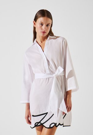 White shirt dress made of lightweight fabric, featuring a button-down front, 3/4 sleeves, and a tied waist, with a black logo at the hem.