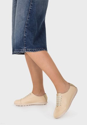 Piernas con vaqueros cortos y zapatillas beige de cuero sin cordones con cordones elásticos sobre un fondo claro.