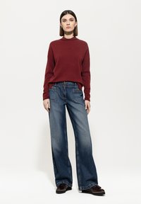 Bordeauxroter Strickpullover mit hohem Kragen, kombiniert mit weit geschnittenen blauen Jeans aus Denim, die über Vorder- und Gesäßtaschen sowie eine ausgewaschene Optik verfügen.