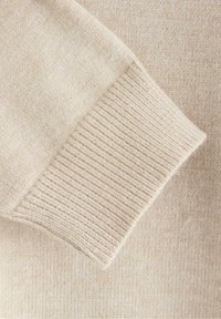 Läheltä kuvattu beige neulepusero, jossa on ribbausvarsi ja tasainen pinta. Kankaan sävyissä on hienovaraisia vaihteluita.