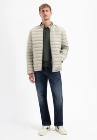 Beige gewatteerde pufferjack met een ronde kraag, gedragen over een groene languarige shirt en donkerblauwe jeans, aangevuld met witte sneakers.