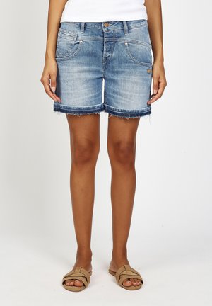 Woman wearing light blue denim shorts with frayed hems and tan slide sandals standing against a plain background.