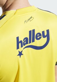 Žlté športové tričko s modrým textom "halley" a dizajnom hviezdy. Na zadnej strane sa nachádza podpis. Vyrobené z hladkého, priedušného materiálu.