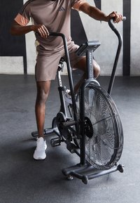 Bici de aire con un gran ventilador negro, marco metálico y manillares ajustables. Usuario con ropa deportiva marrón y zapatillas blancas. Entorno de fitness.