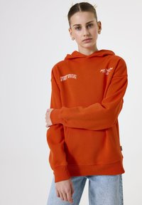 Young woman with blonde hair in an orange hoodie with "New York" text, adjusting her sleeve, standing against a plain light background.