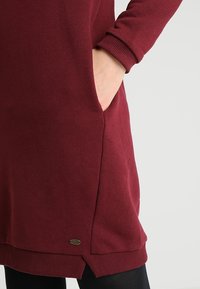 Vestido burdeos con puños acanalados, bolsillos laterales y un pequeño detalle de logotipo metálico en el dobladillo. Textura suave y corte relajado.