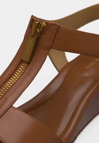 Καφέ δερμάτινο σανδάλι με χρυσό φερμουάρ στην κορυφή και σταυρωτό λουράκι πάνω από το πόδι, με τακούνι σε σφήνα και επένδυση στην εσωτερική σόλα.