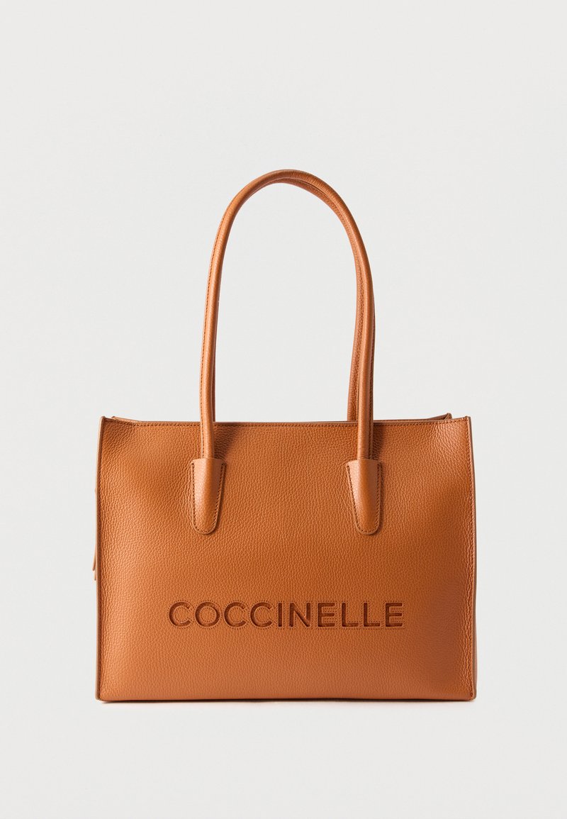 Τσάντα ώμου από δέρμα σε αποχρώσεις ταμπά με δύο χειρολαβές και το λογότυπο "COCCINELLE" ανάγλυφο στην πρόσοψη, σε λευκό αφαιρετικό φόντο.
