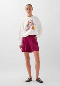 Hanorac alb cu un design grafic colorat, purtat împreună cu pantaloni scurți magenta. Modelul poartă pantofi slip-on negri, cu mâinile în buzunare. Fundal simplu.