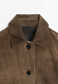 Hnědá semišová bunda s výrazným límcem, zapínáním na jeden černý knoflík a kontrastním prošitím podél okrajů. Hladká textura.