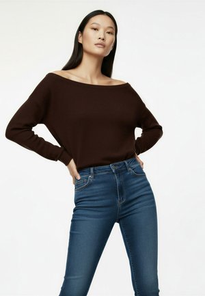 Femme debout, les mains sur les hanches, portant un haut marron foncé sans épaules et un jean bleu taille haute sur fond blanc.