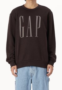 Tmavě hnědá mikina se šedým logem "GAP", žebrovaným výstřihem do kulata a manžetami. Na sobě má světle modré denimové šortky. Měkká textura materiálu.