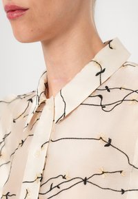 Gros plan d'une personne portant une chemise crème avec des motifs floraux brodés noirs et beiges et un col pointu.