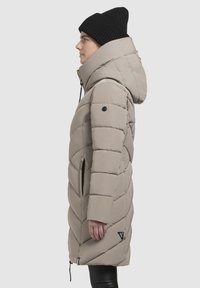 Beige insulated puffer coat with a cinched hood, diamond pattern, side pockets, and black logo accent. Worn with a black knitted beanie.