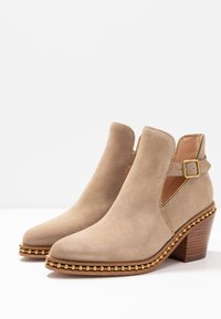 Beżowe zamszowe botki sięgające kostki z ozdobnymi złotymi ćwiekami, wyciętymi bokami, paskiem z klamrą oraz warstwowanymi, drewnianymi obcasami na białej powierzchni.