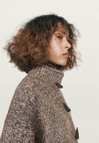 Hnědý pletený svetr s vysokým límcem, vyrobený z texturované tkaniny a tmavých knoflíků. Design má volný střih a jedinečný vzor.