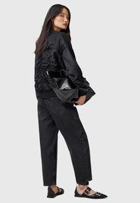 Giacca bomber nera in satin con schiena arricciata, indossata con jeans neri larghi e scarpe nere piatte con accessori dorati, mentre tiene una borsa nera lucida.