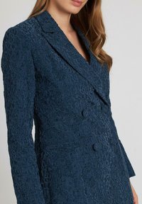 Blazer de doble botonadura en azul marino con patrón de encaje texturizado, solapas con muesca y dos botones decorativos en el frente.