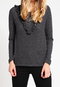 Dunkelgrauer Pullover mit strukturiertem Strick, langen Ärmeln und einem Rüschendetail am Ausschnitt. Getragen mit schwarzen Hosen, die das Design des Oberteils unterstreichen.