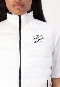 Baltas pūkų liemuo su horizontaliu siuvinėjimo raštu, aukšta apykakle ir juodu užtrauktuku. Kairėje pusėje yra juodas firminis logotipas.