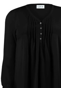 Svart blus med V-ringning, med vertikala plisseringar fram, sex knappar och långa, något rynkade ärmar. Tillverkad av ett slätt tyg.