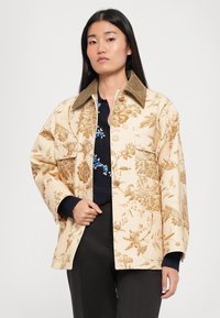 Ziedu raksta krēms jakas ar korduroja apkakli, ar priekšējiem kabatām un brīvu piegriezumu, uzvilkta virs tumši zila džempera.