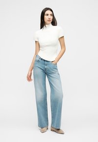 Γυναίκα με μακριά σκουρόχρωμα μαλλιά, φορώντας λευκό ριμπ μπλουζάκι με κοντά μανίκια, ανοιχτό μπλε τζιν παντελόνι με φαρδιά μπατζάκια και μπεζ παπούτσια, που στέκεται μπροστά σε λευκό φόντο.