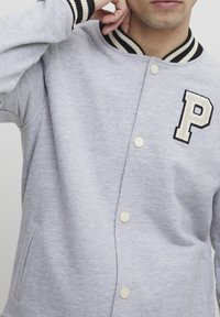 Grå sweatshirt med knappeåpning, ribbestrikket krage med svart og hvite striper, og et stort brodert bokstav "P" i svart og krem.