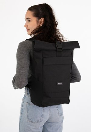 Giovane donna che indossa un maglione grigio e jeans chiari porta uno zaino nero grande con chiusura a rotolo, tasca anteriore e fibbia.