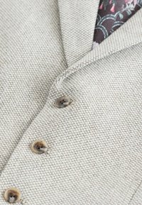 Helle beige, strukturierte Stoff mit subtilen Muster, ausgestattet mit einem Reverskragen und drei braunen Knöpfen, die ein floral gemustertes Innenfutter enthüllen.