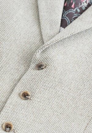 Ljusbeige strukturerad tyg med ett subtilt mönster, med en notch-krage och tre bruna knappar, som avslöjar ett blommigt foder.